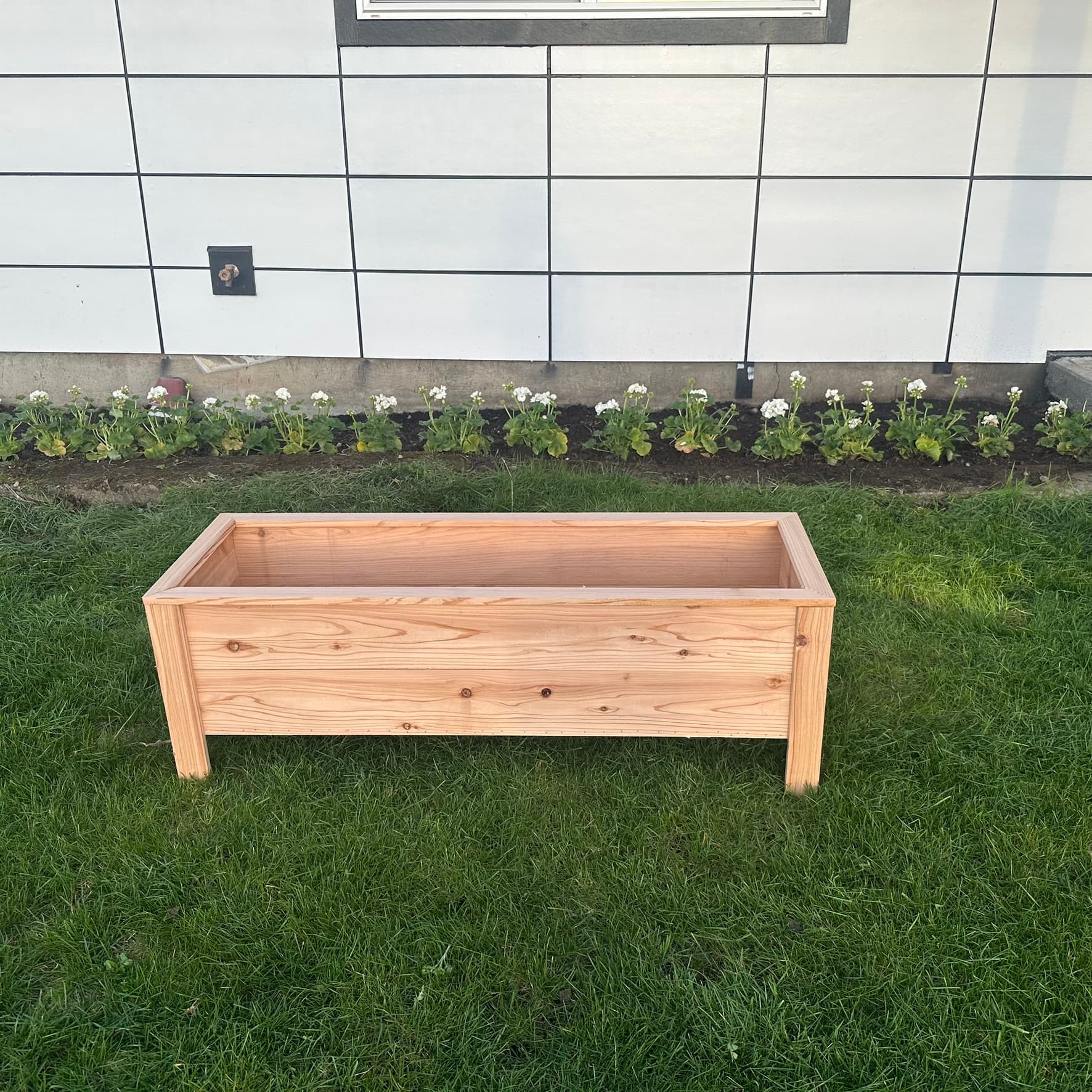 Compact cedar planter box with short legs, front view with flower bed in background